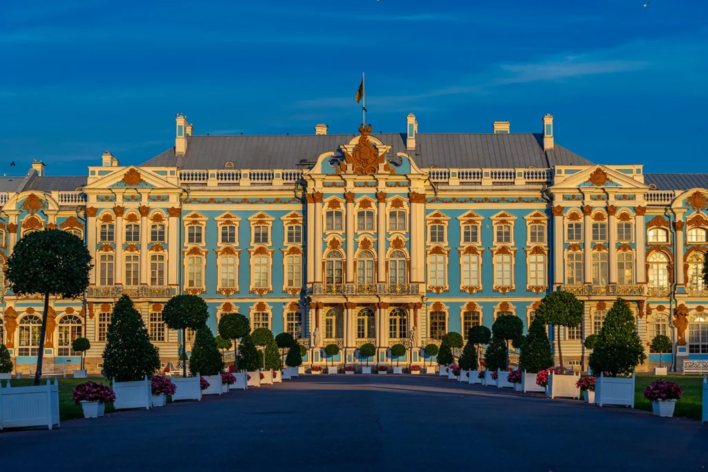The magnificent blue and gold facade of the Catherine Palace illuminated by sunlight, with a pathway of manicured trees.