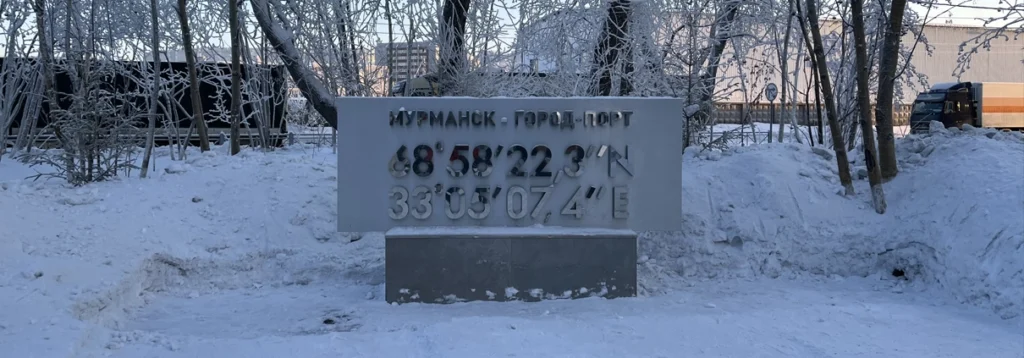 The official coordinates sign and geographic marker for Murmansk, Russia, the largest city above the Arctic Circle, covered in heavy winter snow and surrounded by frosted trees.